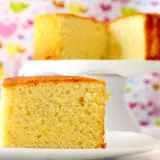 Un trozo de bizcocho de yogur esponjoso sobre un plato blanco. Detrás, otro bizcocho en un soporte, con un fondo de corazones de colores.