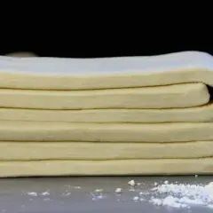 Masa de tarta de hojaldre doblada en capas, lista para hornear. Un rodillo de madera y harina espolvoreada al fondo.