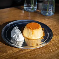 Delicioso flan de yogur con caramelo líquido y una porción de nata montada, servido en un plato metálico sobre una mesa de madera.