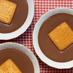 Tres cuencos de natillas de chocolate, cada uno con una galleta cuadrada encima, sobre un mantel de cuadros rojos y blancos.