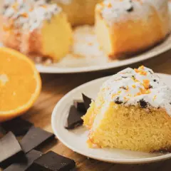 Un delicioso bizcocho de chocolate y naranja, espolvoreado con azúcar glas y ralladura de naranja, acompañado de trozos de chocolate negro y una naranja fresca.