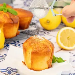 Magdalenas de limón doradas, espolvoreadas con azúcar glas. Limones frescos y menta decoran la escena.