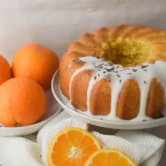 Bizcocho de naranja y yogur, recién horneado y decorado con glaseado blanco y virutas de chocolate. Naranjas frescas y rodajas de naranja completan la escena.