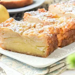 Porción de tarta de manzana con capas finas, espolvoreada con azúcar glas. Dos manzanas amarillas al lado.