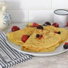 Deliciosos crepes de avena doblados y cubiertos con bayas congeladas y frescas. Servidos en un plato blanco con cubiertos y una jarra de cerámica al fondo.