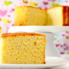 Un trozo de bizcocho de yogur esponjoso sobre un plato blanco. Detrás, otro bizcocho en un soporte, con un fondo de corazones de colores.
