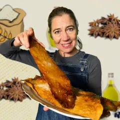 Mujer sonriente muestra una coca de vidre dorada y crujiente, con ingredientes como harina, anís estrellado y aceite de oliva de fondo.