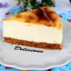 Rebanada de new york cheesecake con base crujiente y relleno cremoso, servida en plato blanco con la palabra "Delicious".