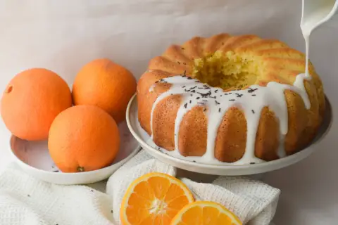 Bizcocho de naranja y yogur, recién horneado y decorado con glaseado blanco y virutas de chocolate. Naranjas frescas y rodajas de naranja completan la escena.