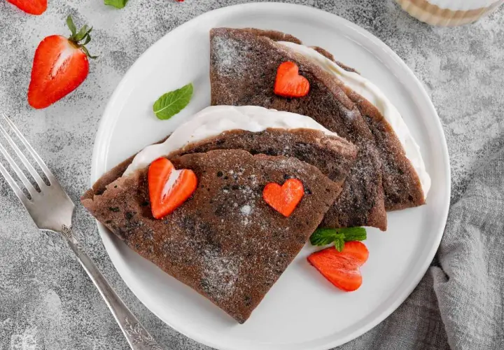 Deliciosos crepes de chocolate rellenos de crema, decorados con fresas en forma de corazón y espolvoreados con azúcar glas.