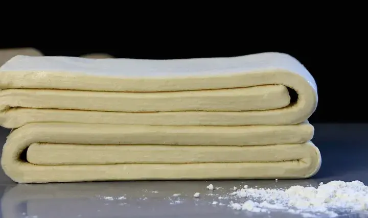 Masa de tarta de hojaldre doblada en capas, lista para hornear. Un rodillo de madera y harina espolvoreada al fondo.