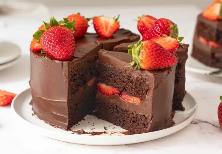 Deliciosa tarta de fresas y chocolate, cubierta de ganache y decorada con fresas frescas. Un trozo revela capas de bizcocho de chocolate y fresas.