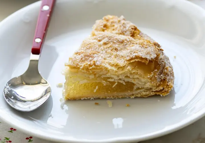 Un trozo de pastel cordobés, hojaldrado y cubierto de azúcar, espera ser disfrutado junto a una cuchara en un plato blanco.