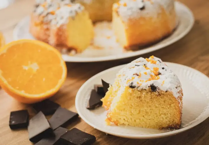 Un delicioso bizcocho de chocolate y naranja, espolvoreado con azúcar glas y ralladura de naranja, acompañado de trozos de chocolate negro y una naranja fresca.