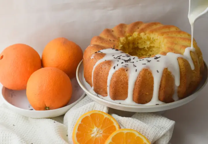 Bizcocho de naranja y yogur, recién horneado y decorado con glaseado blanco y virutas de chocolate. Naranjas frescas y rodajas de naranja completan la escena.