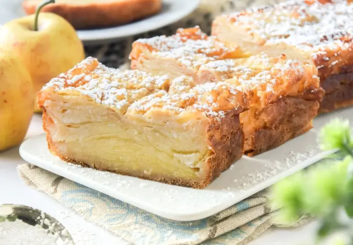 Porción de tarta de manzana con capas finas, espolvoreada con azúcar glas. Dos manzanas amarillas al lado.