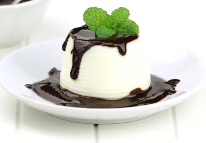 Deliciosa panna cotta de chocolate blanco, cubierta con salsa de chocolate y decorada con hojas de menta fresca.