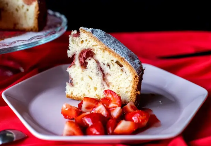Rebanada de bizcocho de fresa esponjoso, espolvoreado con azúcar glas, servido con fresas frescas picadas en un plato rosa.