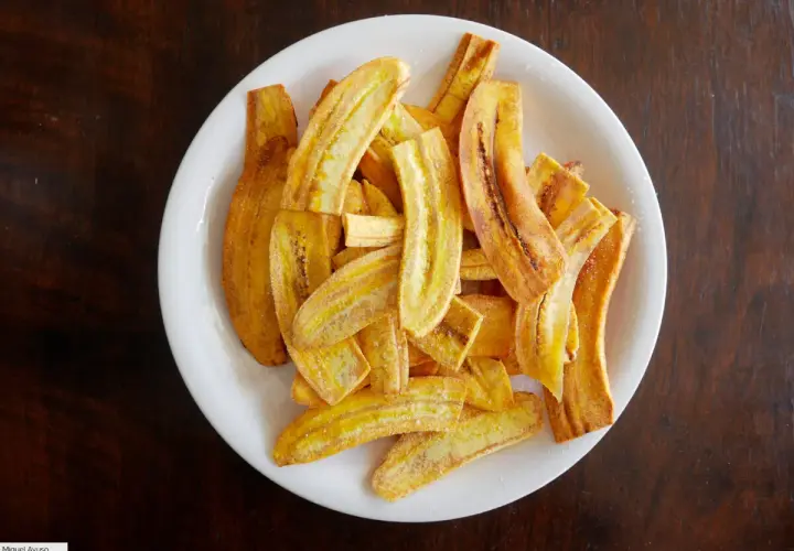 Un plato blanco lleno de crujientes rodajas de plátano frito, espolvoreadas con azúcar y canela.