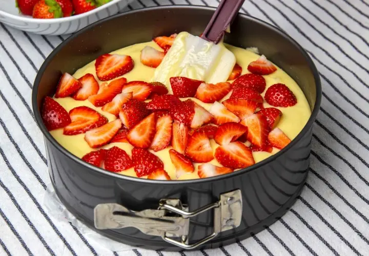 Bizcocho de yogur de fresa en molde, con fresas cortadas esparcidas sobre la masa y una espátula.