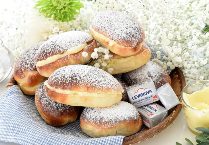 Bollos de mantequilla cubiertos de azúcar glas, rellenos de crema, junto a levadura y flores blancas.