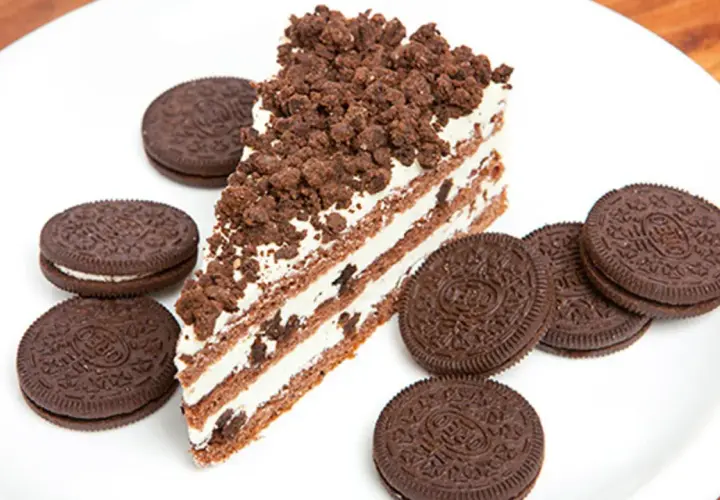 Rebanada de bizcocho de oreo con capas de crema y galleta triturada, rodeada de galletas oreo enteras en un plato blanco.