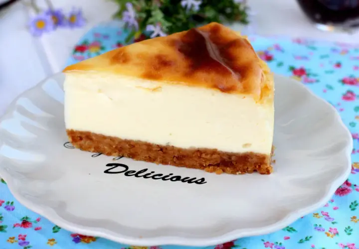 Rebanada de new york cheesecake con base crujiente y relleno cremoso, servida en plato blanco con la palabra "Delicious".