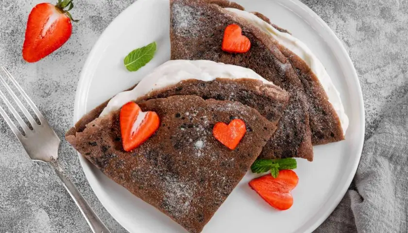 Deliciosos crepes de chocolate rellenos de crema, decorados con fresas en forma de corazón y espolvoreados con azúcar glas.
