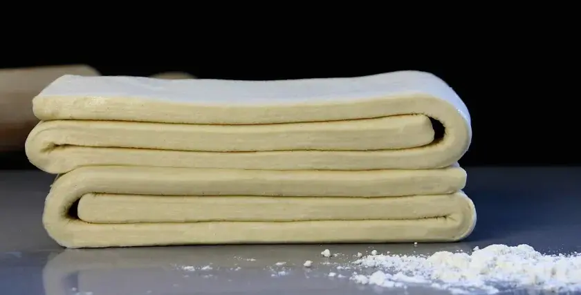 Masa de tarta de hojaldre doblada en capas, lista para hornear. Un rodillo de madera y harina espolvoreada al fondo.