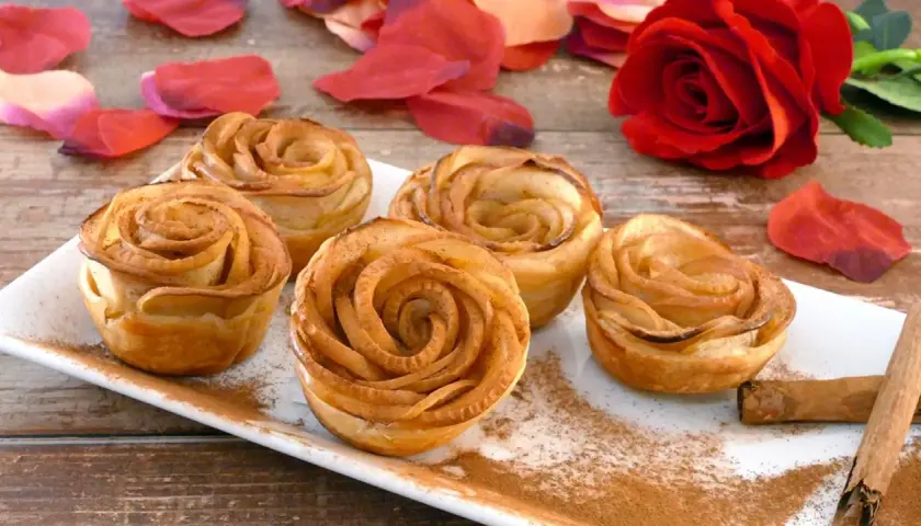 Deliciosas rosas de manzana horneadas, espolvoreadas con canela, junto a pétalos rojos y una rosa natural.