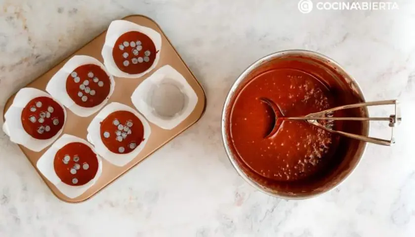 Molde con magdalenas de chocolate listas para hornear y un bol con masa de chocolate y cucharón.