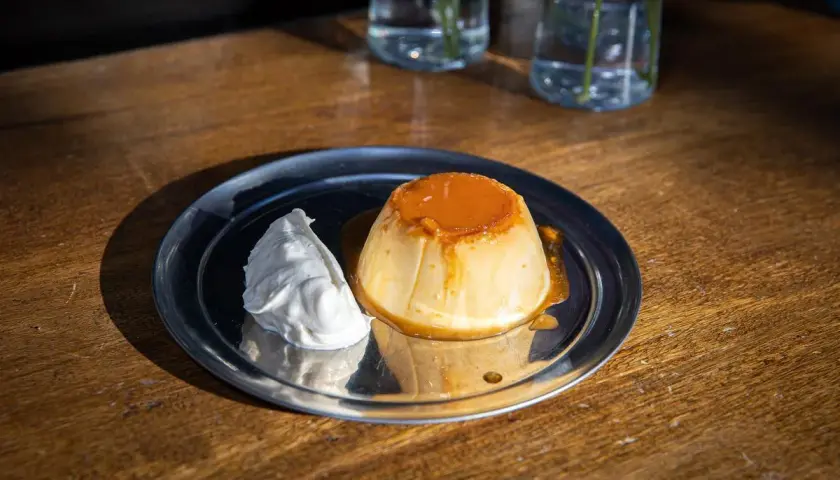 Delicioso flan de yogur con caramelo líquido y una porción de nata montada, servido en un plato metálico sobre una mesa de madera.