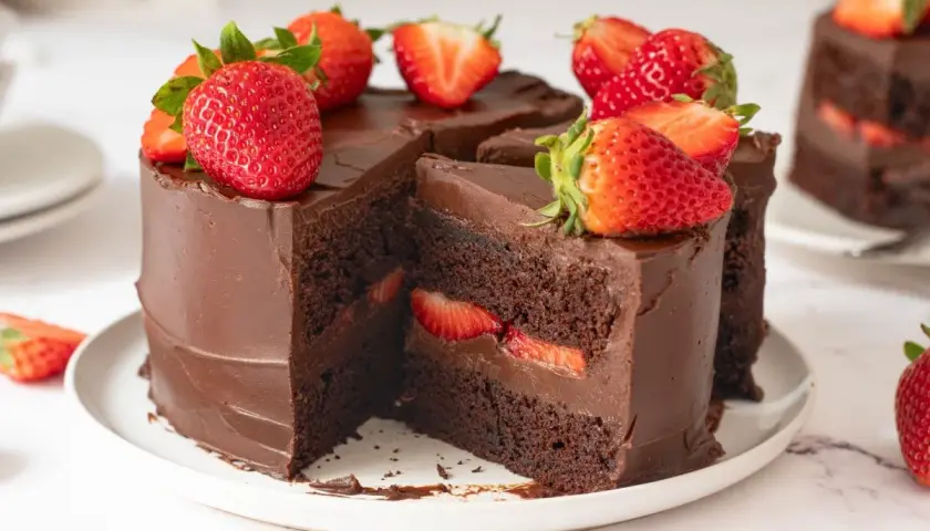Deliciosa tarta de fresas y chocolate, cubierta de ganache y decorada con fresas frescas. Un trozo revela capas de bizcocho de chocolate y fresas.