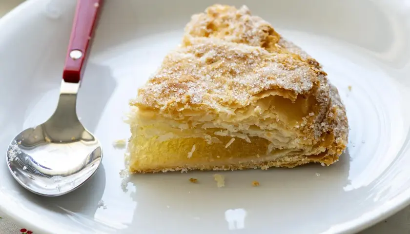 Un trozo de pastel cordobés, hojaldrado y cubierto de azúcar, espera ser disfrutado junto a una cuchara en un plato blanco.