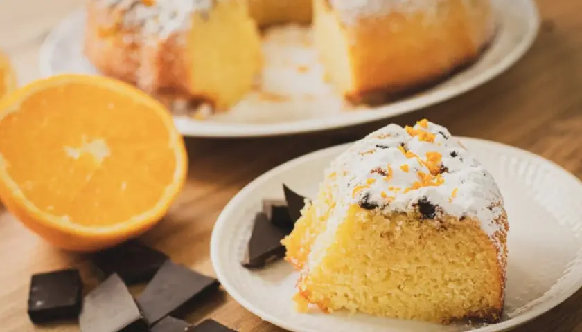 Un delicioso bizcocho de chocolate y naranja, espolvoreado con azúcar glas y ralladura de naranja, acompañado de trozos de chocolate negro y una naranja fresca.