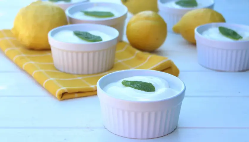 Delicioso mousse de limón mambo en ramequines blancos, adornado con hojas de menta. Limones frescos y una servilleta amarilla completan la escena.