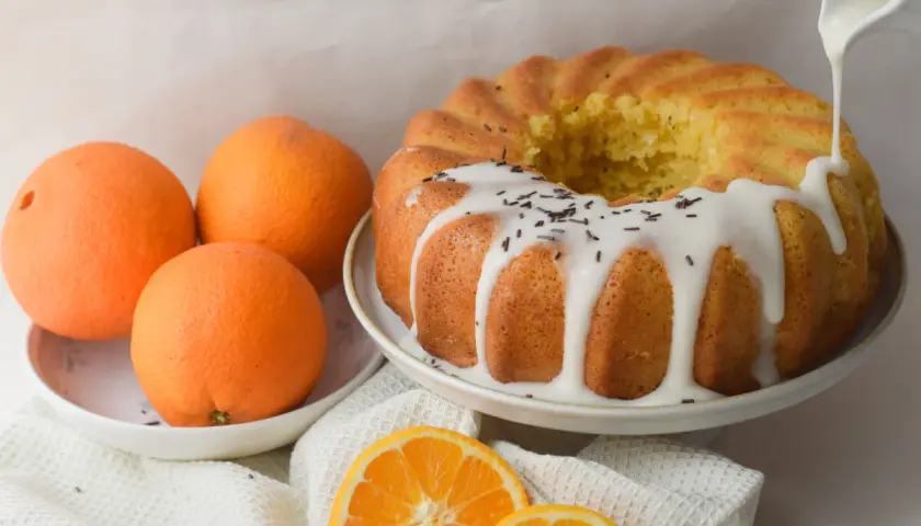 Bizcocho de naranja y yogur, recién horneado y decorado con glaseado blanco y virutas de chocolate. Naranjas frescas y rodajas de naranja completan la escena.