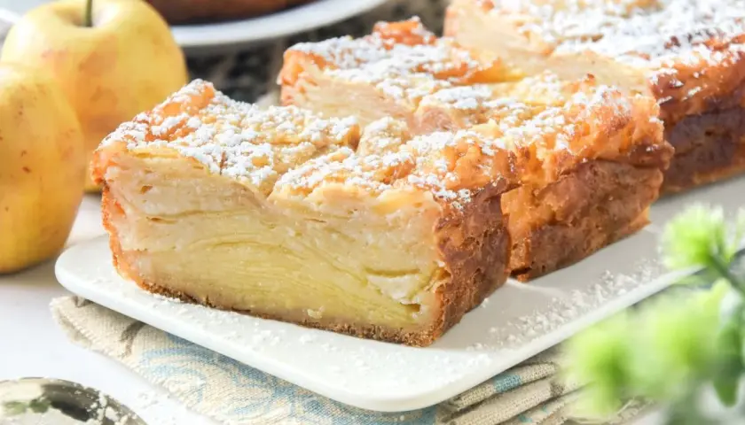 Porción de tarta de manzana con capas finas, espolvoreada con azúcar glas. Dos manzanas amarillas al lado.