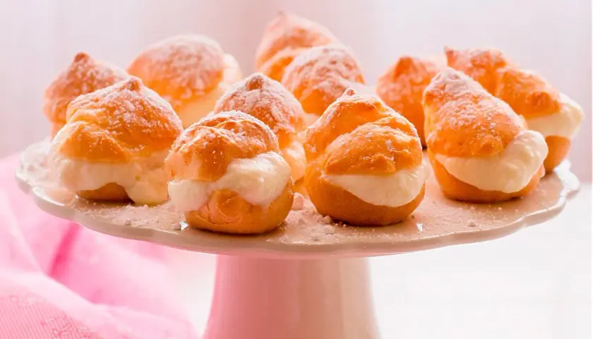 Deliciosos profiteroles de nata, espolvoreados con azúcar glas, listos para ser disfrutados en una bandeja blanca.