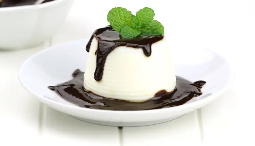 Deliciosa panna cotta de chocolate blanco, cubierta con salsa de chocolate y decorada con hojas de menta fresca.