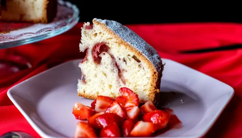 Rebanada de bizcocho de fresa esponjoso, espolvoreado con azúcar glas, servido con fresas frescas picadas en un plato rosa.