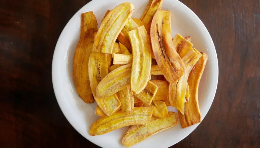 Un plato blanco lleno de crujientes rodajas de plátano frito, espolvoreadas con azúcar y canela.