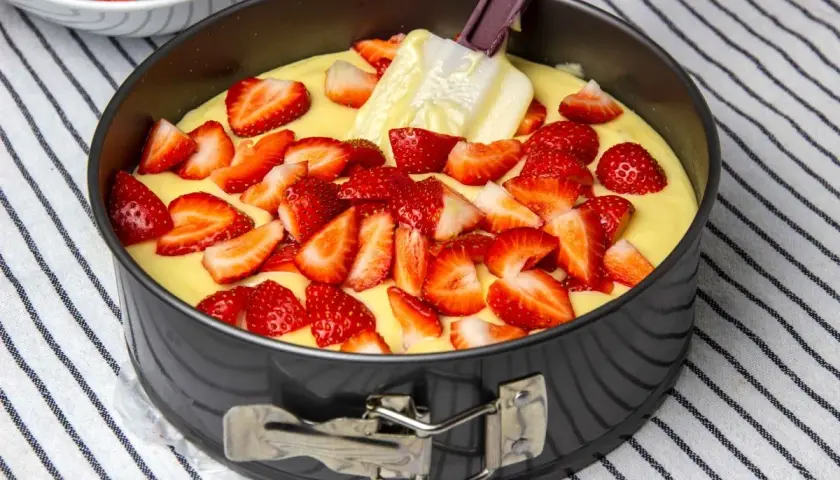 Bizcocho de yogur de fresa en molde, con fresas cortadas esparcidas sobre la masa y una espátula.