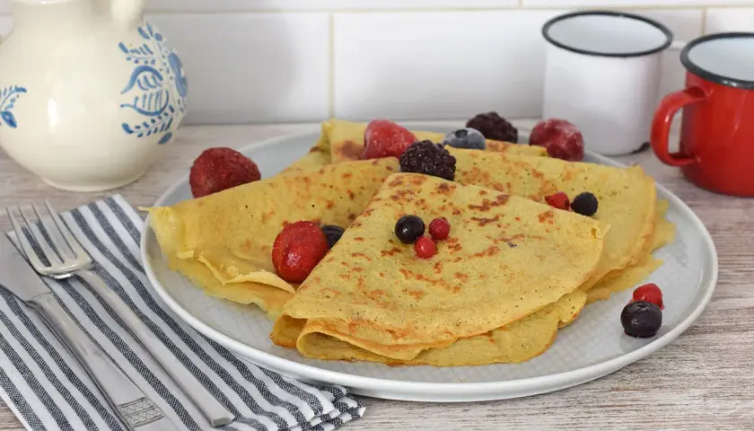 Deliciosos crepes de avena doblados y cubiertos con bayas congeladas y frescas. Servidos en un plato blanco con cubiertos y una jarra de cerámica al fondo.