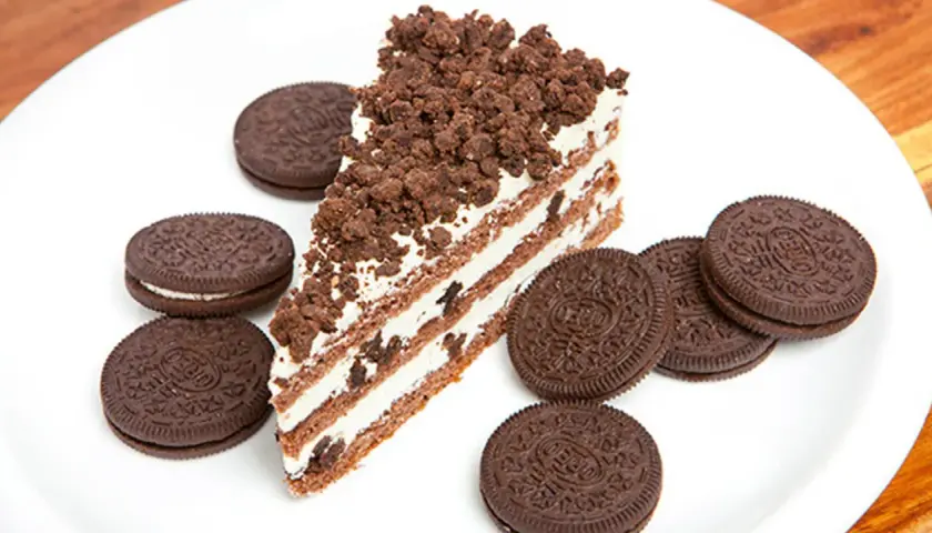Rebanada de bizcocho de oreo con capas de crema y galleta triturada, rodeada de galletas oreo enteras en un plato blanco.