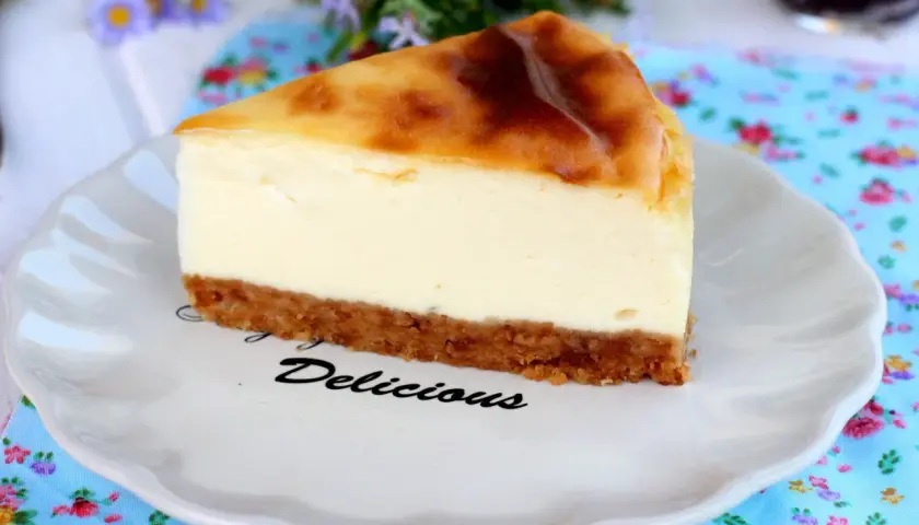 Rebanada de new york cheesecake con base crujiente y relleno cremoso, servida en plato blanco con la palabra "Delicious".