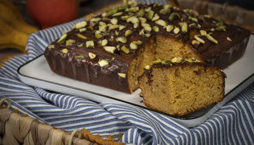 Bizcocho con harina de espelta, cubierto de chocolate y pistachos. Una rebanada muestra su interior esponjoso.