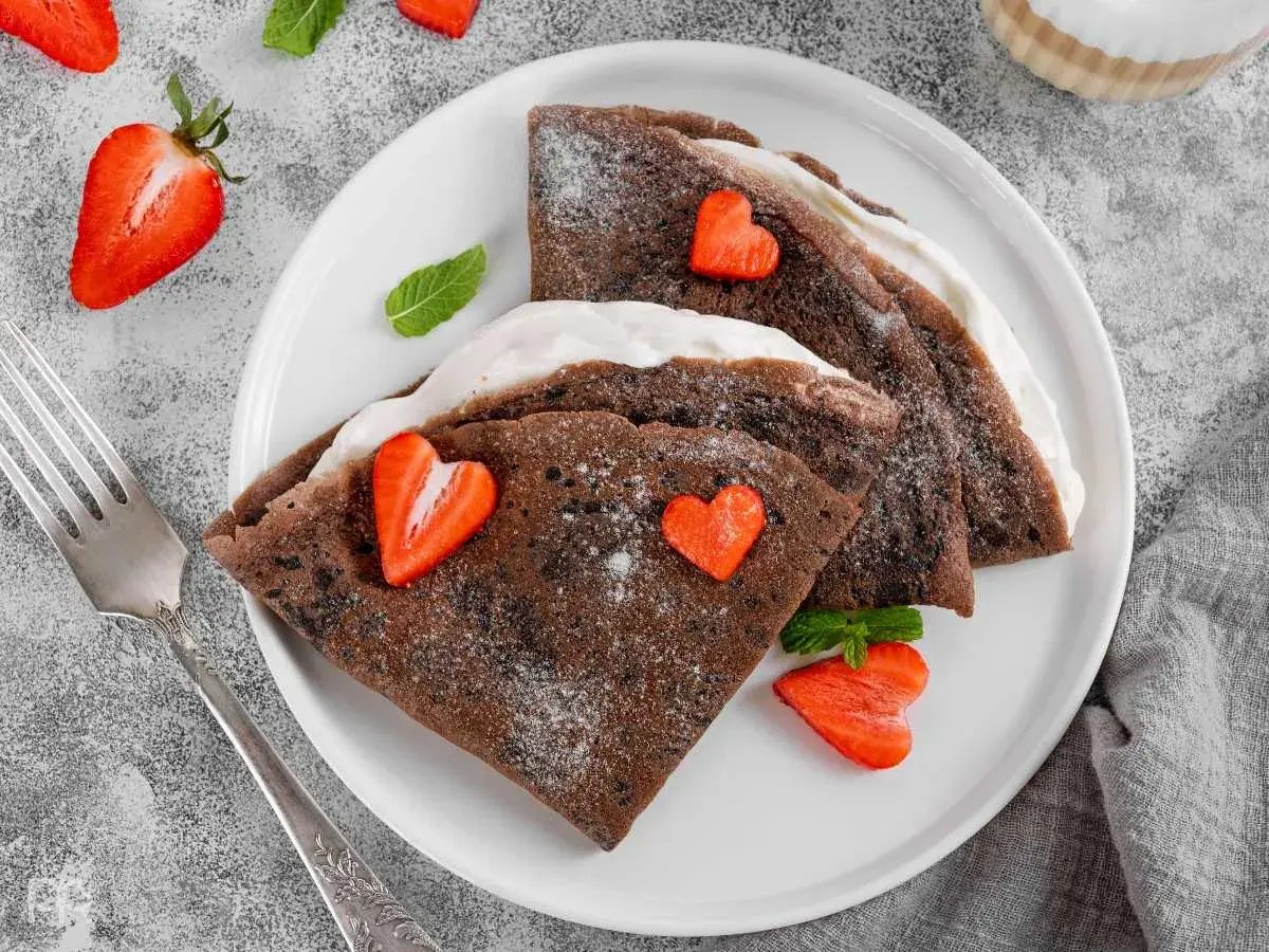 Deliciosos crepes de chocolate rellenos de crema, decorados con fresas en forma de corazón y espolvoreados con azúcar glas.