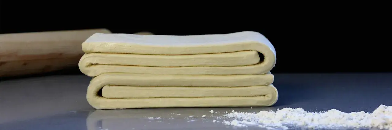 Masa de tarta de hojaldre doblada en capas, lista para hornear. Un rodillo de madera y harina espolvoreada al fondo.