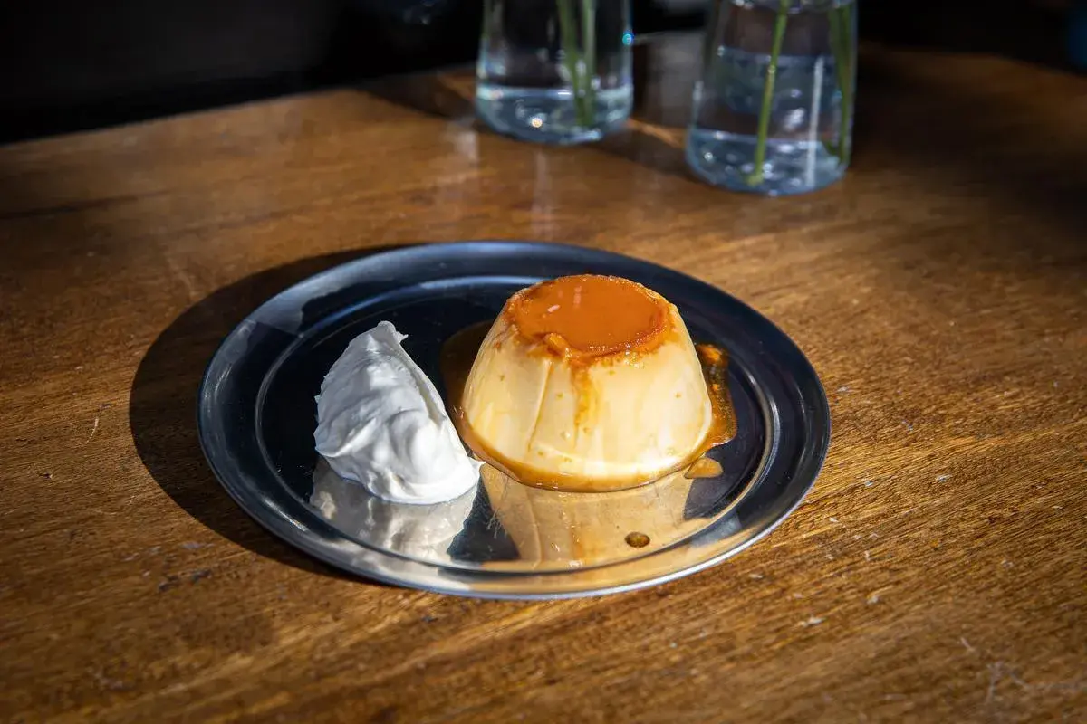 Delicioso flan de yogur con caramelo líquido y una porción de nata montada, servido en un plato metálico sobre una mesa de madera.